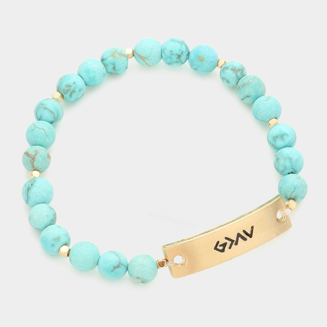 A symbolic bracelet with a metal bar and natural stones, displaying the faith-inspired message "God is Greater Than the Highs and Lows."